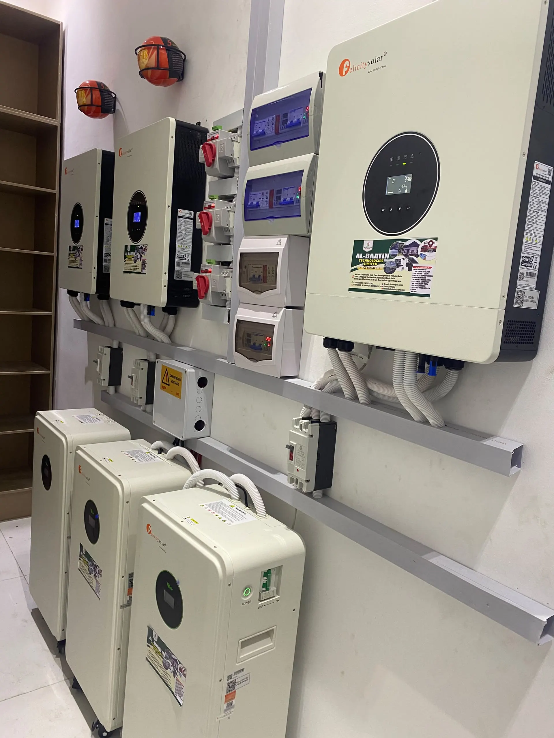Three Felicity hybrid inverters mounted in a row above a rail of breakers and changeover switches, with three floor-standing lithium batteries below and Al-Baatin stickers on each unit.