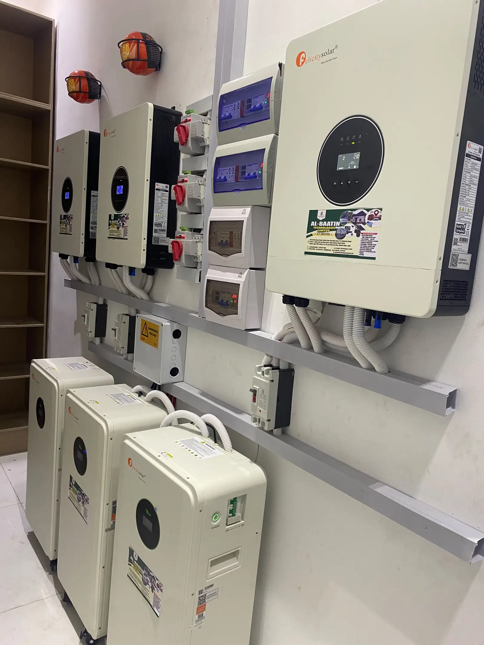 Three Felicity hybrid inverters mounted in a row above three floor-standing lithium batteries, with breaker boxes between them and Al-Baatin stickers on the units.