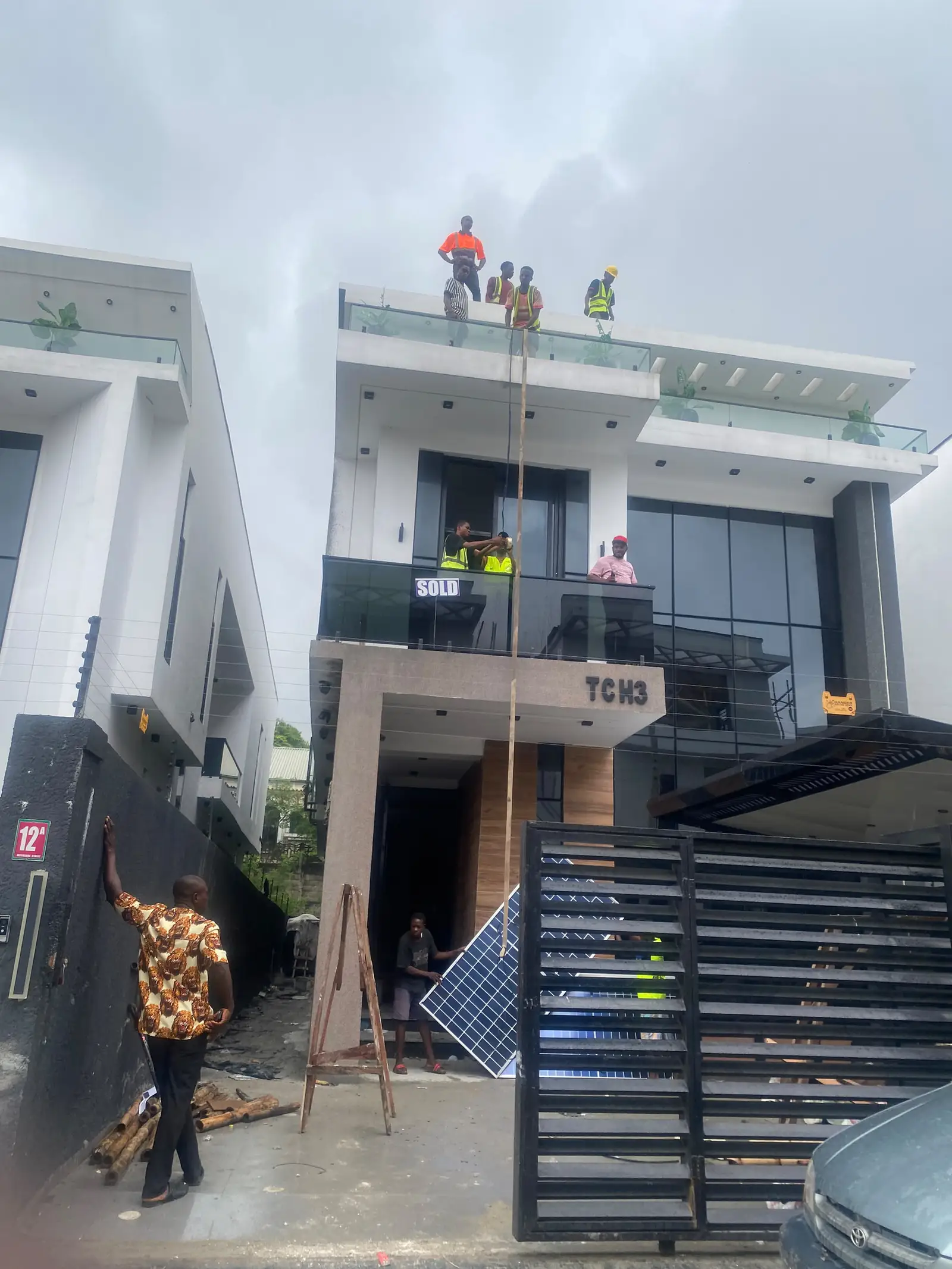 Al-Baatin fitters in hi-vis vests working on the roof terrace and balcony to mount a solar panel on a two-storey white duplex at Daffodil Gardens Estate.