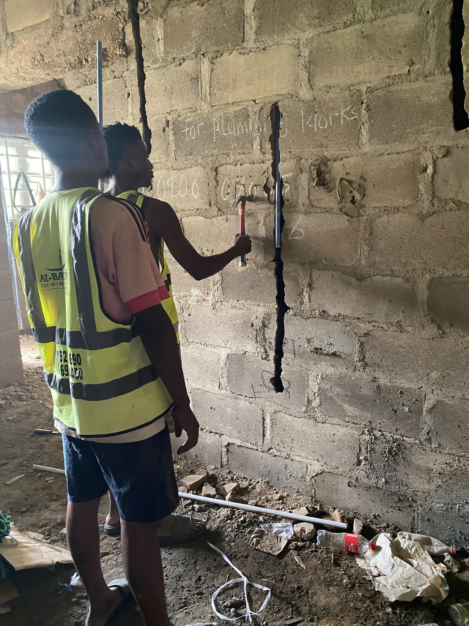 Two Al-Baatin fitters in hi-vis vests cutting a channel down a block wall with a hammer and chisel, the route for the conduit chalked on the blockwork beside them.