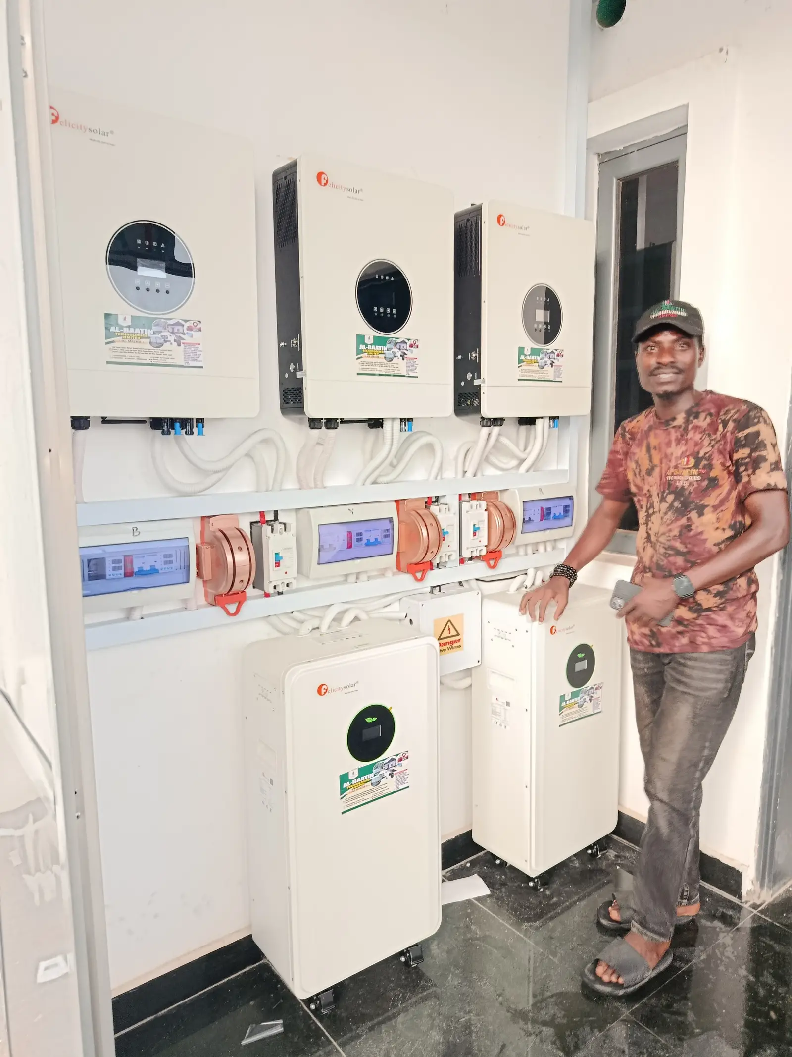 Three Felicity hybrid inverters above a row of breaker boxes, with two floor-standing lithium batteries below and Al-Baatin standing beside them.