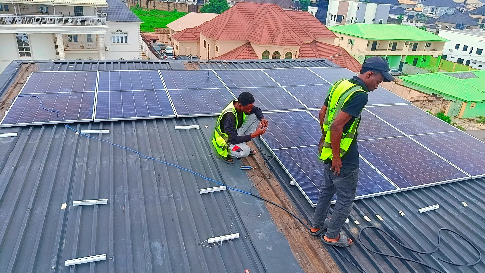 Two Al-Baatin fitters in hi-vis vests fixing solar panels to a dark metal roof, with the rooftops of the estate behind them.