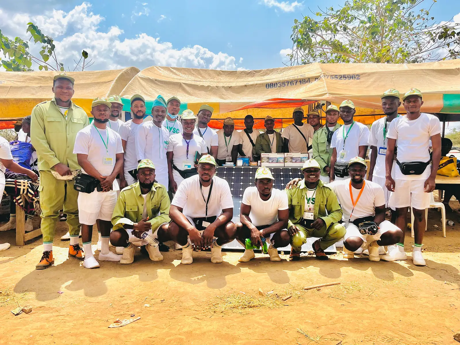 A large group of corps members in NYSC kit crouched and standing around a solar panel at the Al-Baatin training stand in camp.